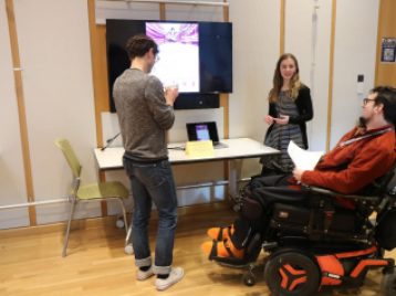 Claire Callen stands beside a large monitor displaying her presentation as she explains her project to two people.